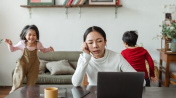 A lady trying to work on her laptop with her two young kids running around the room behind her