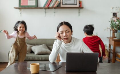 A lady trying to work on her laptop with her two young kids running around the room behind her