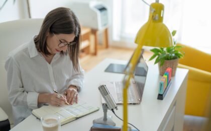 A lady sitting at her desk writing in a book with a laptop and microphone in front of her