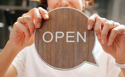 A woman holding a wooden speech bubble with the word OPEN written on it