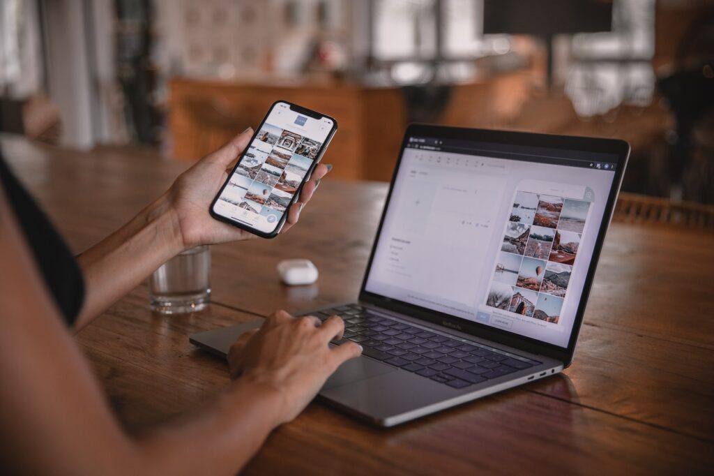 A woman sitting at an open laptop. A mobile phone is in her left hand, displaying a website. Her right hand is on the laptop keyboard, editing the same content that is displayed on the phone.